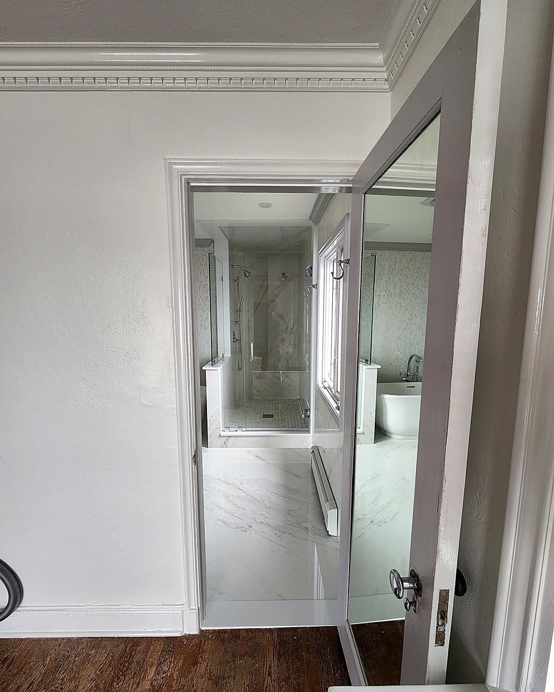 Finished Stamford master bathroom seen from the bedroom doorway, showing marble-look porcelain, glass shower and freestanding tub