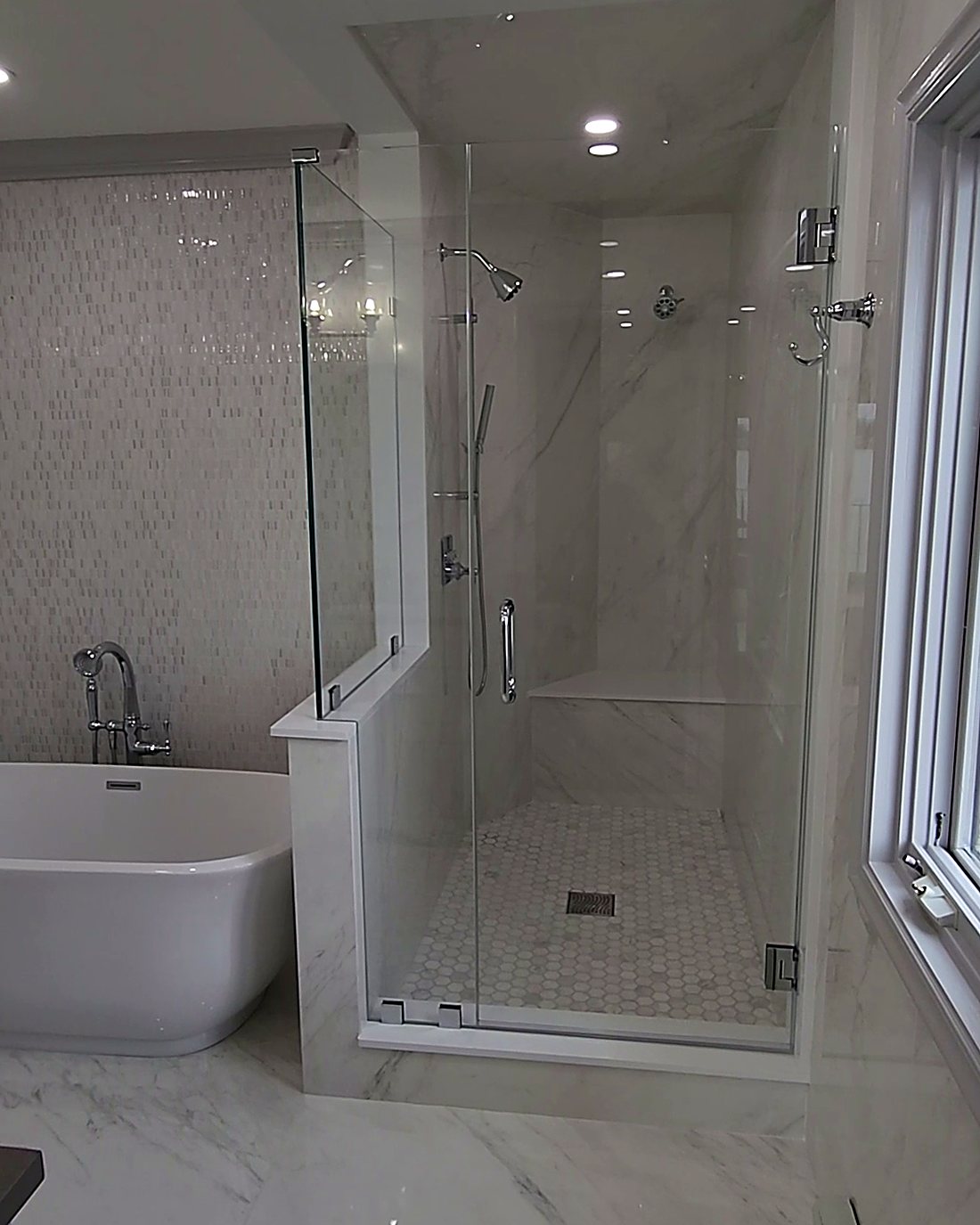 Finished walk-in glass shower with marble-look porcelain, built-in bench and hex mosaic floor beside a freestanding tub