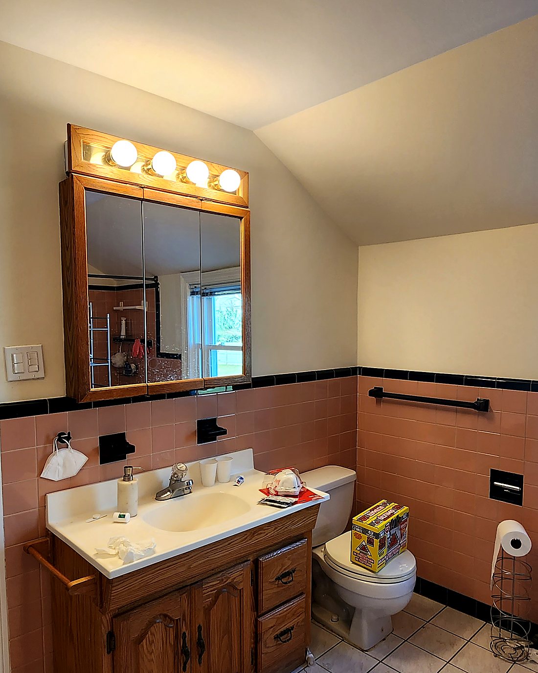 Dated bathroom vanity before remodeling, with an oak vanity, cultured marble sink, Hollywood-bulb mirror and salmon wall tile
